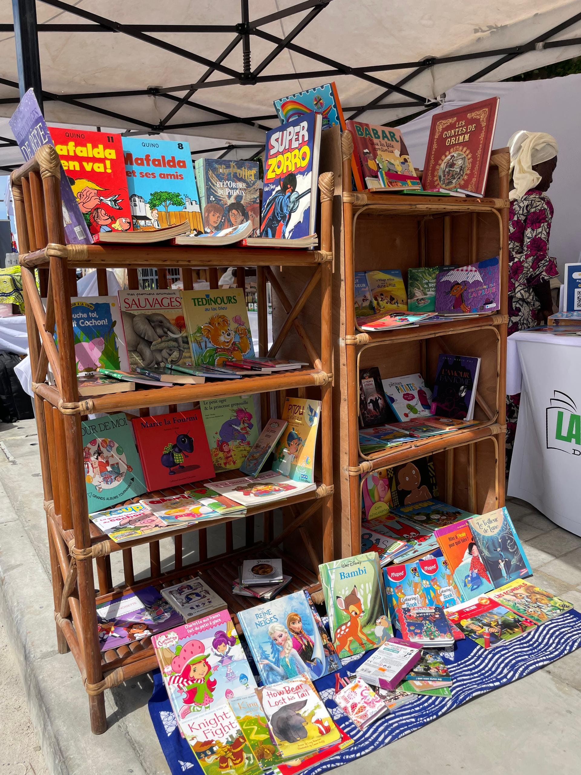 Étagères en bois garnies de livres jeunesse (Mafalda, Bambi, contes) sous une tente d'exposition